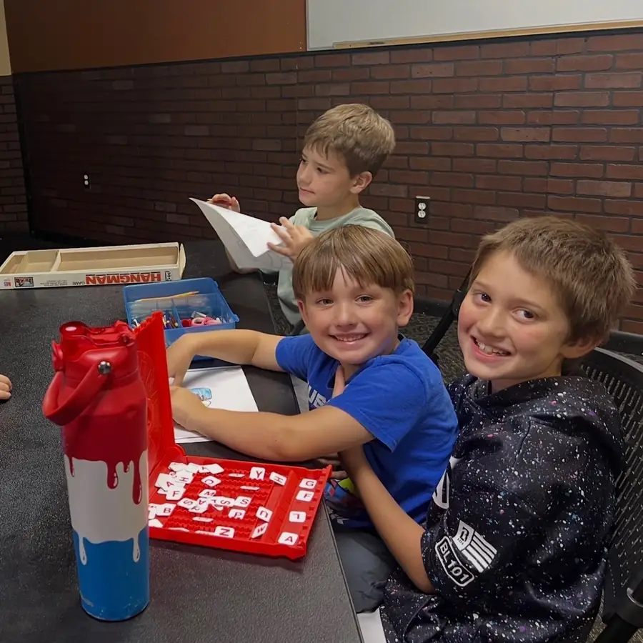 Three boys at a table with a Hangman game and a red water bottle, two of them grinning at the camera