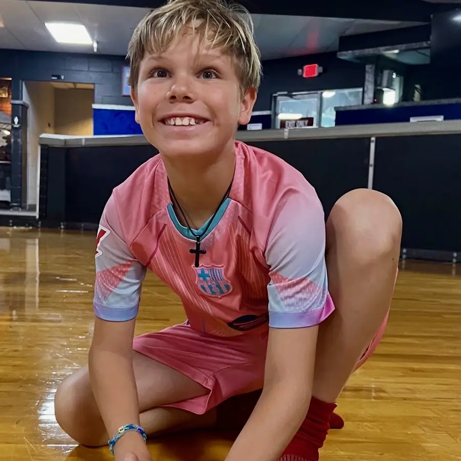 A grinning boy in a pink soccer jersey kneeling on the wooden rink floor at AfterSchool