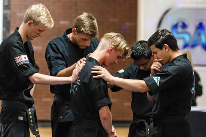 Five students in black uniforms standing in a circle with heads bowed and hands on each other's shoulders, praying together on the dojo floor