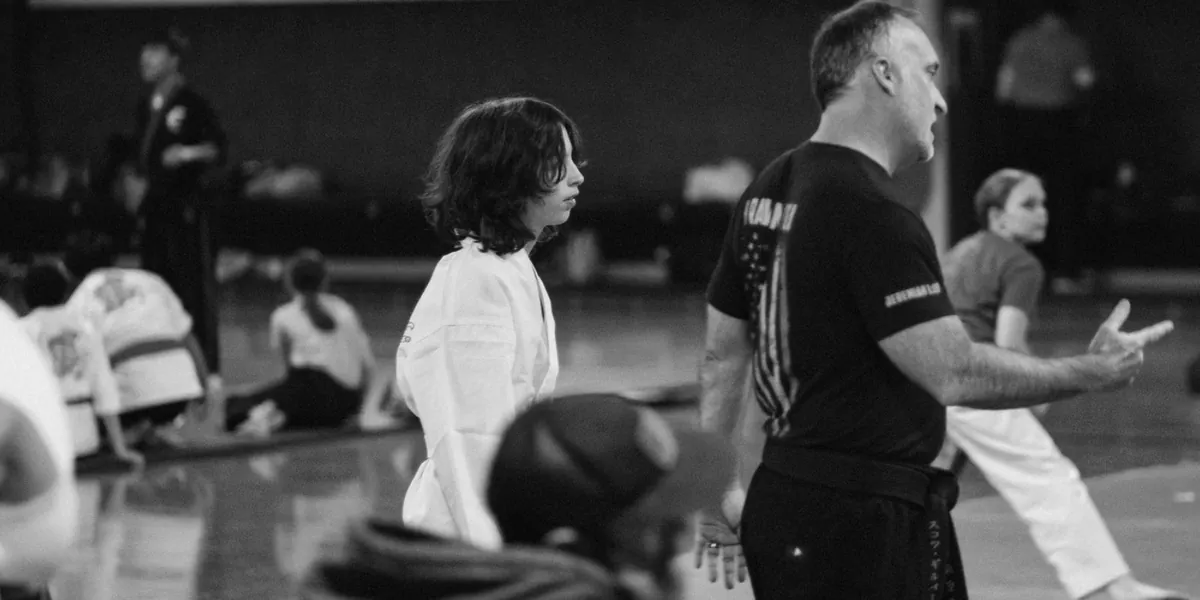 Headmaster Scott Gilbert coaching a class from the floor in profile, a young student in a white gi beside him and students seated on the mat behind