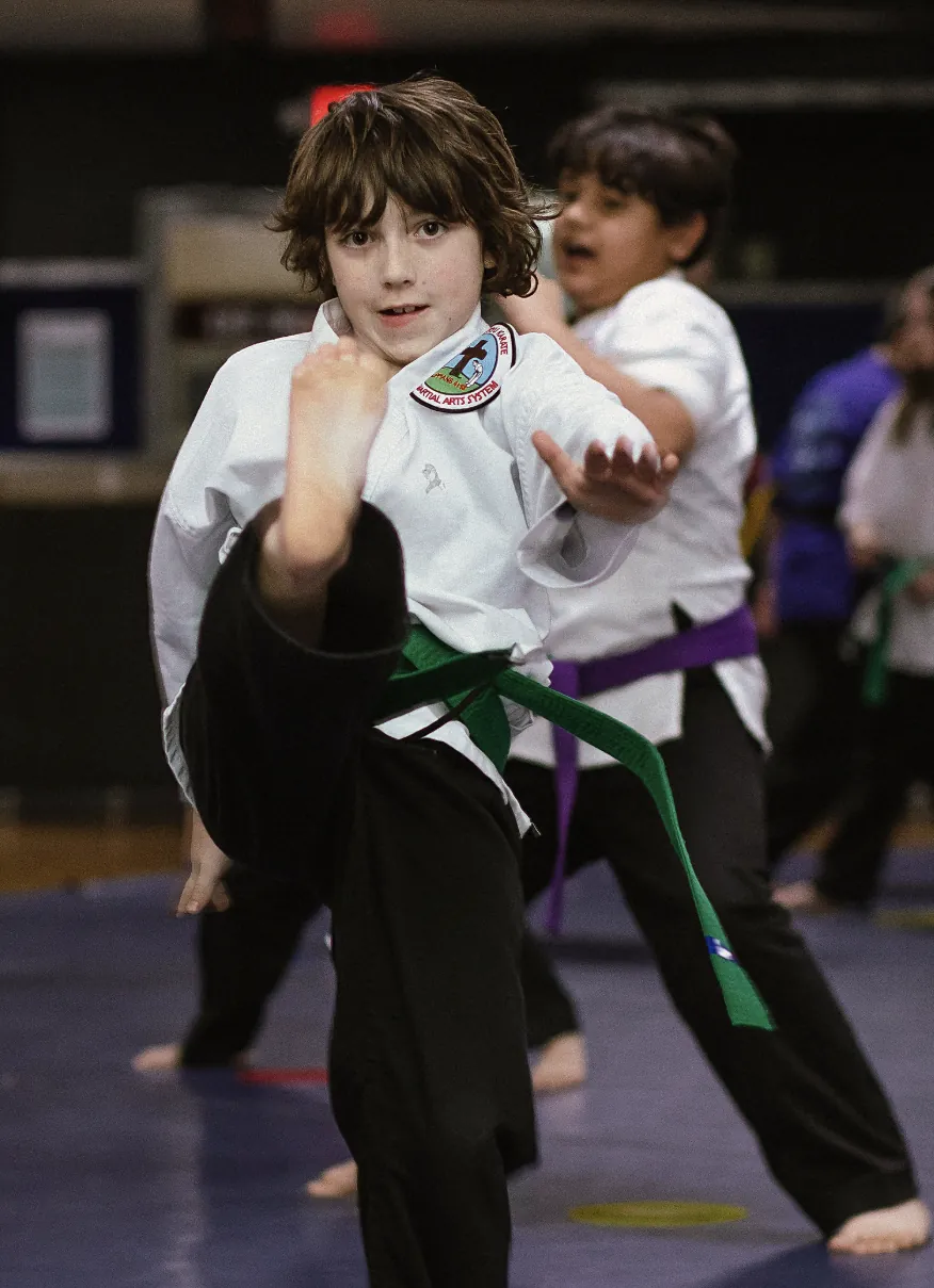 A young student in a white gi and green belt snapping a front kick straight at the camera, with a purple-belt classmate kicking behind him