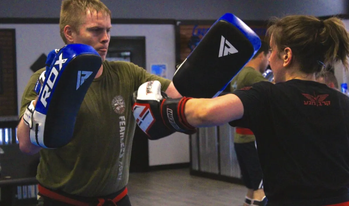 A Krav Maga student in boxing gloves striking focus mitts held up by her training partner