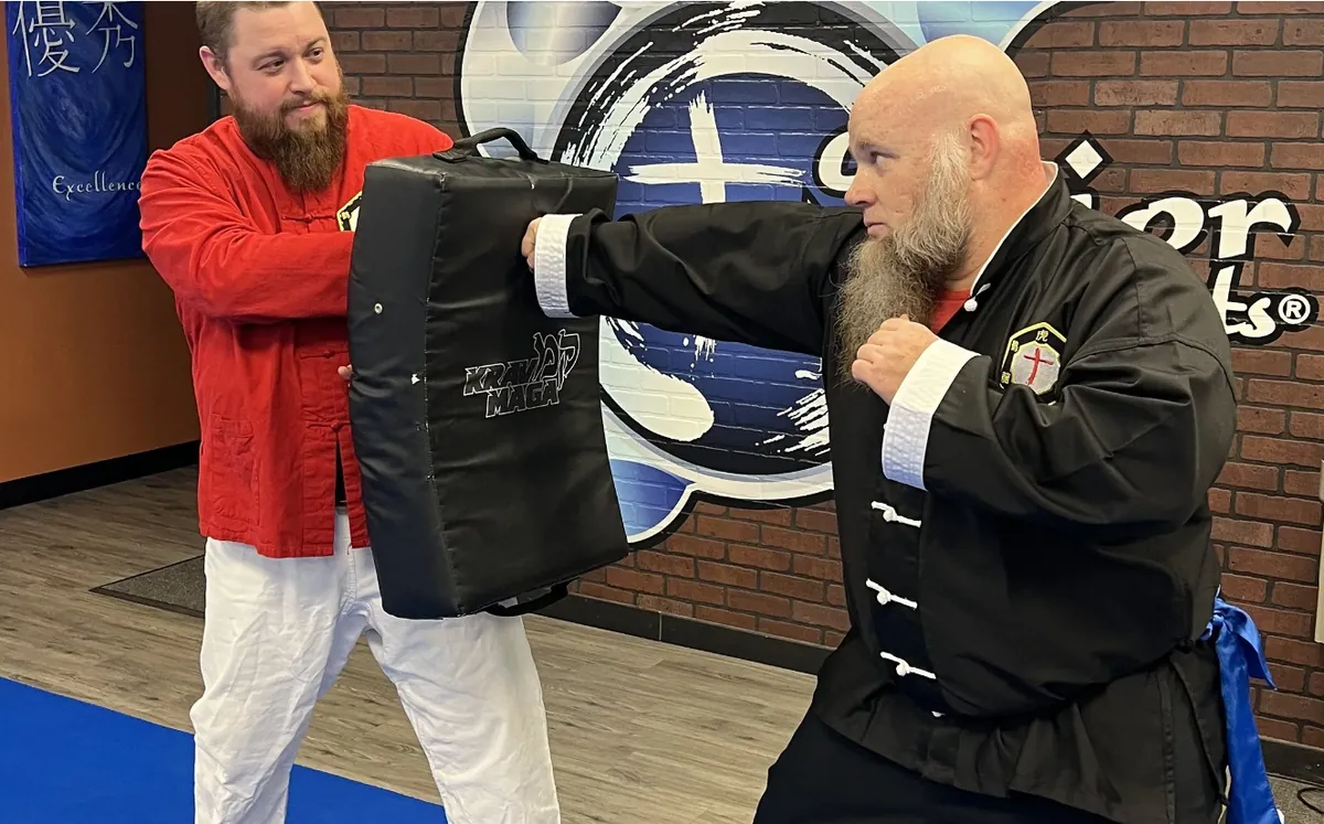 Two Kung Fu students in traditional uniforms, one striking a kick shield held by the other, in front of the school's wall logo