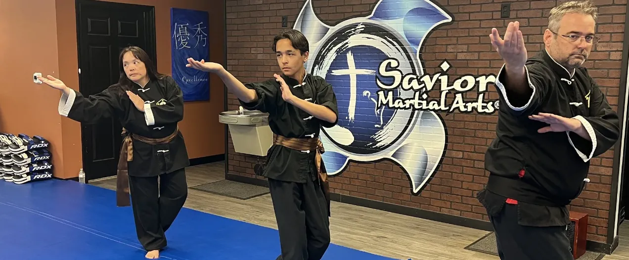 Three Kung Fu students in black uniforms and brown sashes holding an open-hand stance in a line in front of the wall logo