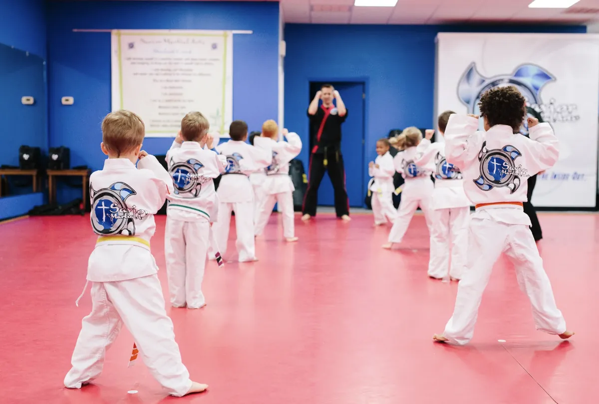 A line of young Dragons in white gis with the school logo on their backs, fists up, following an instructor across the red mats