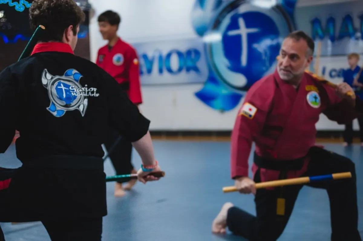 Headmaster Scott Gilbert in his red gi working escrima sticks with a student, the Savior wall logo behind them