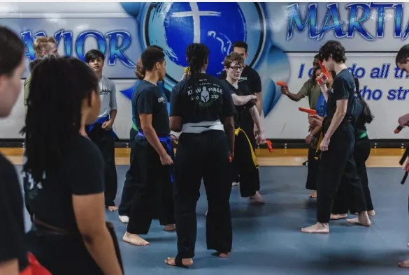 A teen and adult class in black uniforms working a drill with red training knives in front of the wall logo